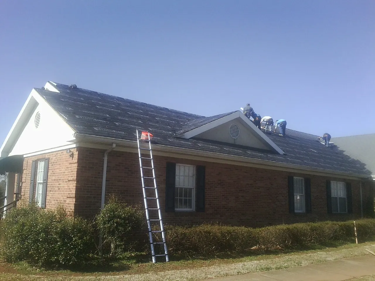 Crew tearing shingles off a brick ranch roof with a ladder set — Emergency Roof Repair work by our crew in Mc Connellsburg