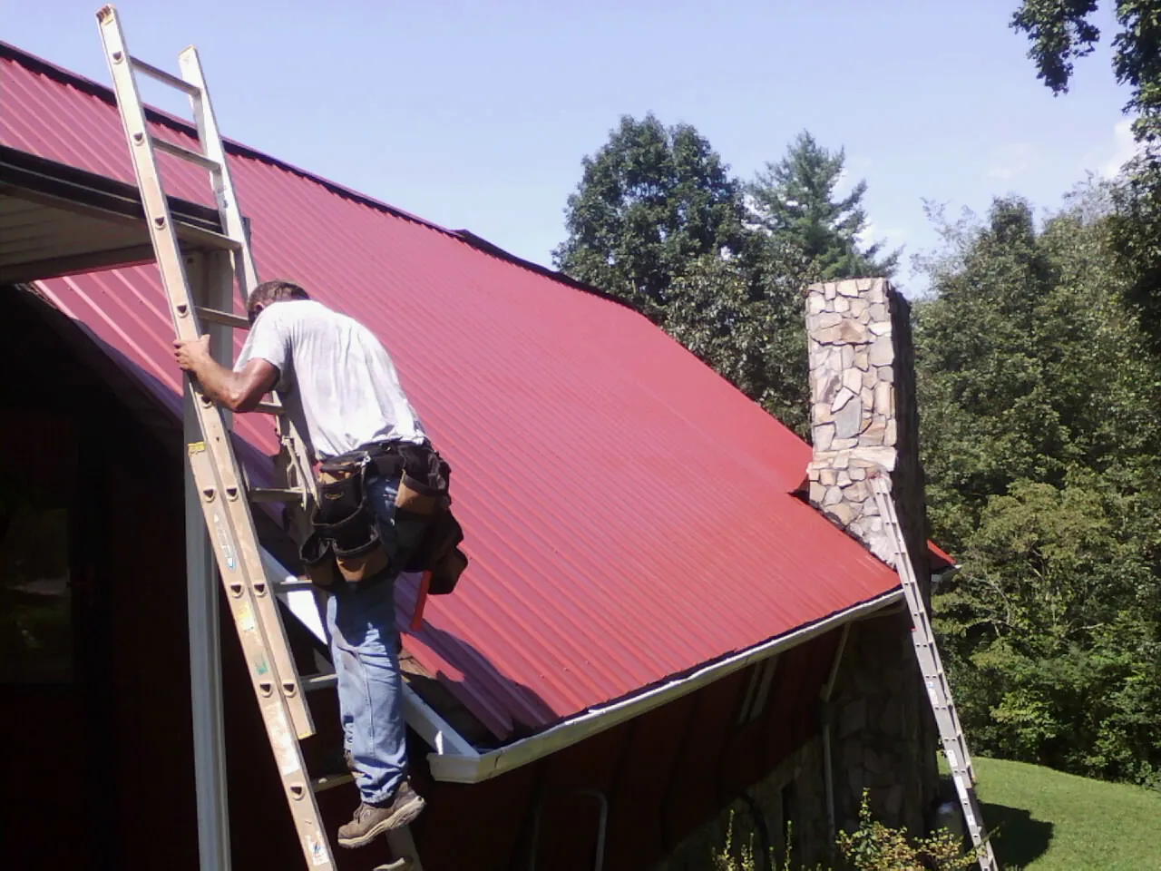 Roofer on a ladder beside a red metal roof and stone chimney — Soffit & Fascia Repair by Roofing Brothers in Mc Connellsburg